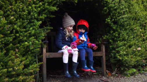 Two children sat on a bench set back into a hedge, looking at a flower head, at Trengwainton Garden, Cornwall